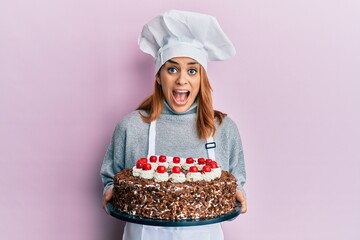 Hispanic young chef woman holding chocolate cake celebrating crazy and amazed for success with open eyes screaming excited.