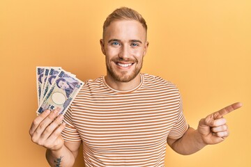 Young caucasian man holding japanese yen banknotes smiling happy pointing with hand and finger to the side