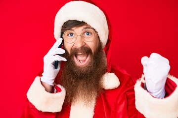 Handsome young red head man with long beard wearing santa claus costume talking on the phone screaming proud, celebrating victory and success very excited with raised arms
