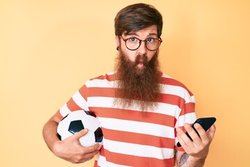 Handsome young red head man with long beard holding football ball looking at smartphone making fish...