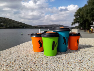 Four multi-colored disposable portable paper coffee cups of different sizes on background of river. Take-Out Coffee.Close-up.