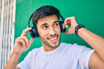 Young latin man smiling happy using headphones leaning on the wall at the city.