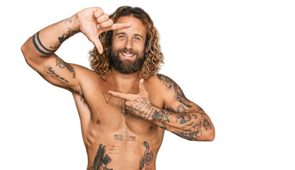 Handsome man with beard and long hair standing shirtless showing tattoos smiling making frame with...