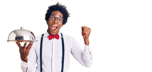 Handsome african american man with afro hair wearing waiter uniform holding silver tray screaming proud, celebrating victory and success very excited with raised arms