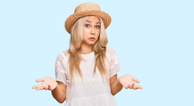 Young blonde girl wearing summer hat clueless and confused expression with arms and hands raised. doubt concept.