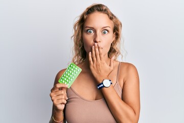 Beautiful caucasian woman holding birth control pills covering mouth with hand, shocked and afraid for mistake. surprised expression