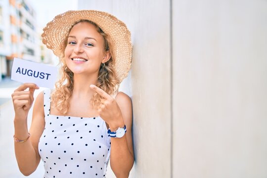 Young beautiful caucasian woman with blond hair smiling happy outdoors on a summer day holding August word