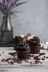 Several muffins or cupcakes with chocolate shaped cream at white table. Festive candle burns on a chocolate cake. There are chocolate chips scattered nearby