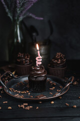 Several muffins or cupcakes with chocolate shaped cream at black table. Festive candle burns on a chocolate cake.