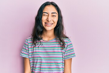 Hispanic teenager girl with dental braces wearing casual clothes winking looking at the camera with sexy expression, cheerful and happy face.