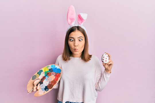 Young beautiful woman wearing cute easter bunny ears holding painting palette and egg making fish face with mouth and squinting eyes, crazy and comical.