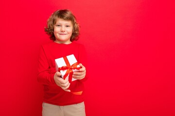 Fototapeta premium Photo shot of joyful smiling blonde curly boy isolated over red background wall wearing red sweater holding gift box and looking at camera