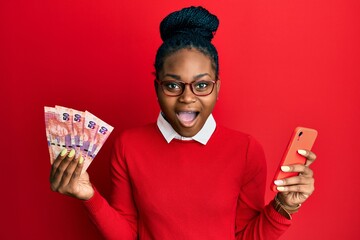 Young african american woman using smartphone holding south africa rand banknotes afraid and...