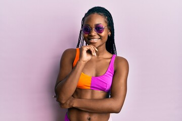 Young african american woman wearing bikini and sunglasses looking confident at the camera with smile with crossed arms and hand raised on chin. thinking positive.