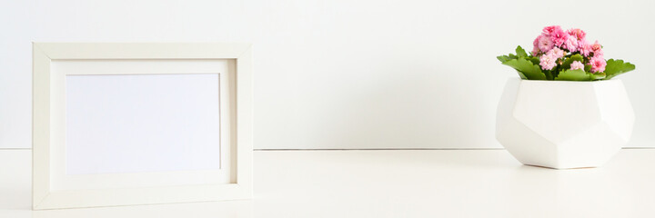 A desk against a white wall. Copy space. Pink flower in a geometric pot. White mockup frame. Banner. Panorama