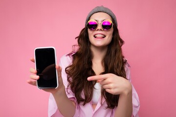 Photo of beautiful smiling young woman good looking wearing casual stylish outfit standing isolated on background with copy space holding smartphone showing phone in hand with empty screen display for