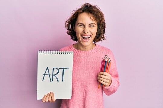 Young brunette woman holding art notebook and colored pencils smiling and laughing hard out loud because funny crazy joke.