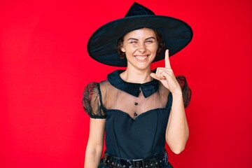 Young hispanic woman wearing witch halloween costume pointing finger up with successful idea. exited and happy. number one.