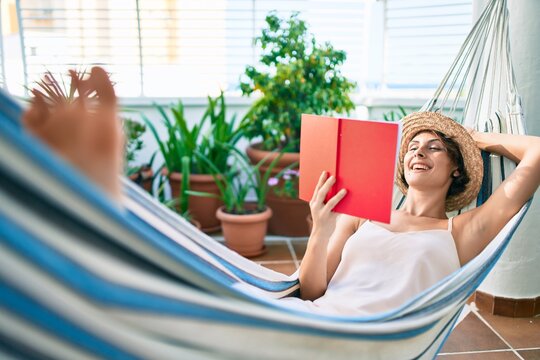 Young beautiful caucasian woman smiling happy resting on a hammock reading a book at home