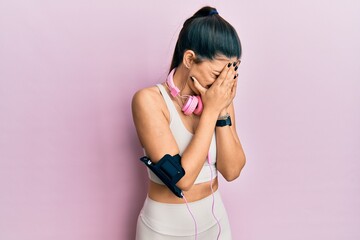 Young hispanic woman wearing gym clothes and using headphones with sad expression covering face with hands while crying. depression concept.