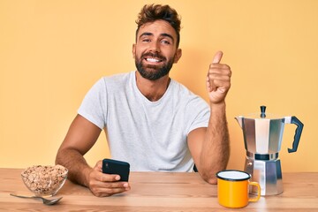 Young hispanic man sitting on the table having breakfast and using smartphone smiling happy and positive, thumb up doing excellent and approval sign