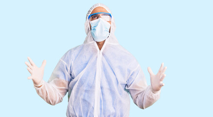 Young hispanic man wearing doctor protection coronavirus uniform and medical mask crazy and mad shouting and yelling with aggressive expression and arms raised. frustration concept.