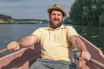 bearded boatman rowing while sitting in a boat, background river and nature