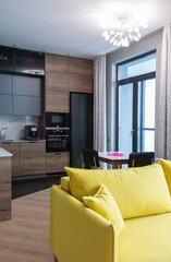 The interior of the studio kitchen is decorated in gray tones with wooden veneers. Kitchen appliances are black. In the foreground is a yellow sofa. Vertical.