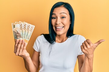 Beautiful hispanic woman holding 500 norwegian krone banknotes pointing thumb up to the side smiling happy with open mouth