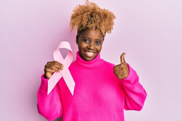 Young african woman with afro hair holding pink cancer ribbon smiling happy and positive, thumb up doing excellent and approval sign