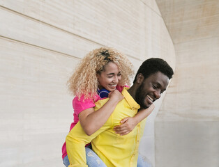 Happy multiracial couple laughing and having fun together with piggyback - Young people enjoy the day in the city - Focus on the face of the man