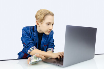 a boy in a jacket works at a laptop and holds banknotes in his hand
