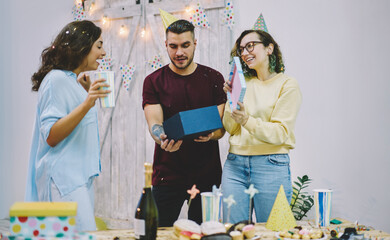Obraz na płótnie Canvas Young hipsters greeting girl on her birthday party opening decorative box with surprise during celebration, happy young woman amazed with gift from best friends satisfied with holiday and festive mood