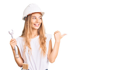 Beautiful caucasian woman with blonde hair wearing hardhat and builder clothes pointing thumb up to the side smiling happy with open mouth