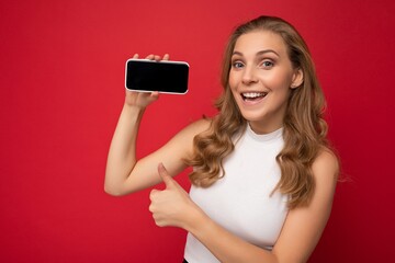 Closeup of happy smiling beautiful young blonde woman wearing white t-shirt isolated on red background with copy space holding smartphone showing phone in hand with empty screen for mockup looking at