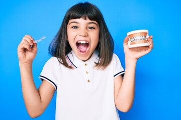 Young little girl with bang holding invisible aligner orthodontic and braces celebrating crazy and amazed for success with open eyes screaming excited.