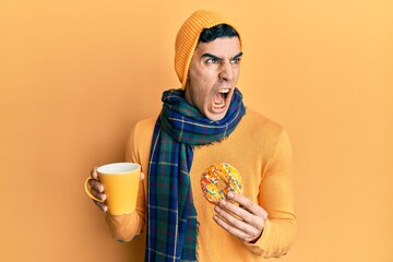 Handsome hispanic man eating doughnut and drinking coffee angry and mad screaming frustrated and...
