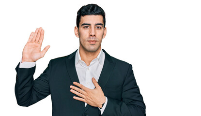 Handsome hispanic man wearing business clothes swearing with hand on chest and open palm, making a...