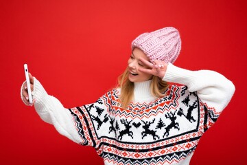 Photo of pretty positive young blonde woman wearing warm knitted hat and winter warm sweater standing isolated over red background using mobile phone taking selfie photo looking at device display and