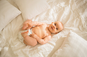 funny little baby girl in a diaper lying on the bed and holding her legs with her hands