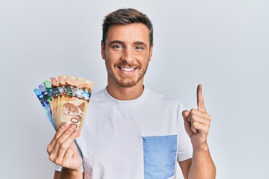 Handsome caucasian man holding canadian dollars smiling with an idea or question pointing finger with happy face, number one