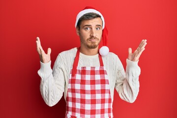 Naklejka premium Handsome caucasian man wearing baker apron and christmas hat clueless and confused with open arms, no idea and doubtful face.