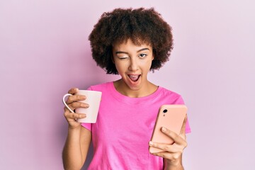 Young hispanic girl using smartphone and drinking a cup of coffee winking looking at the camera...