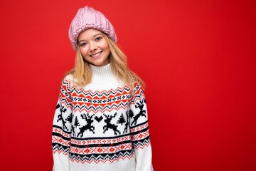 Photo of pretty positive smiling young blonde woman isolated over red background wall wearing winter sweater and pink hat looking at camera and having fun