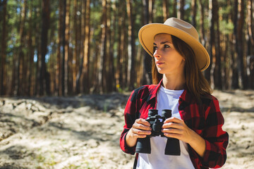 Woman in hat and red plaid shirt holding binoculars in the forest