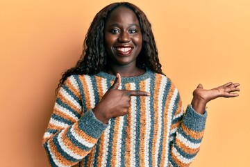 African young woman wearing wool winter sweater amazed and smiling to the camera while presenting with hand and pointing with finger.