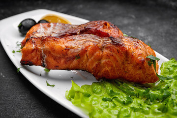 Two baked grilled pieces of salmon fish on a white plate with lettuce, olives and lemon on a dark background