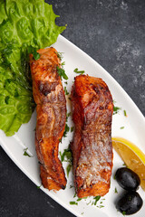 Two baked grilled pieces of salmon fish on a white plate with lettuce, olives and lemon on a dark background