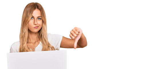 Naklejka premium Beautiful blonde young woman holding blank empty banner with angry face, negative sign showing dislike with thumbs down, rejection concept