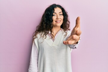 Young brunette woman with curly hair wearing casual clothes smiling friendly offering handshake as greeting and welcoming. successful business.